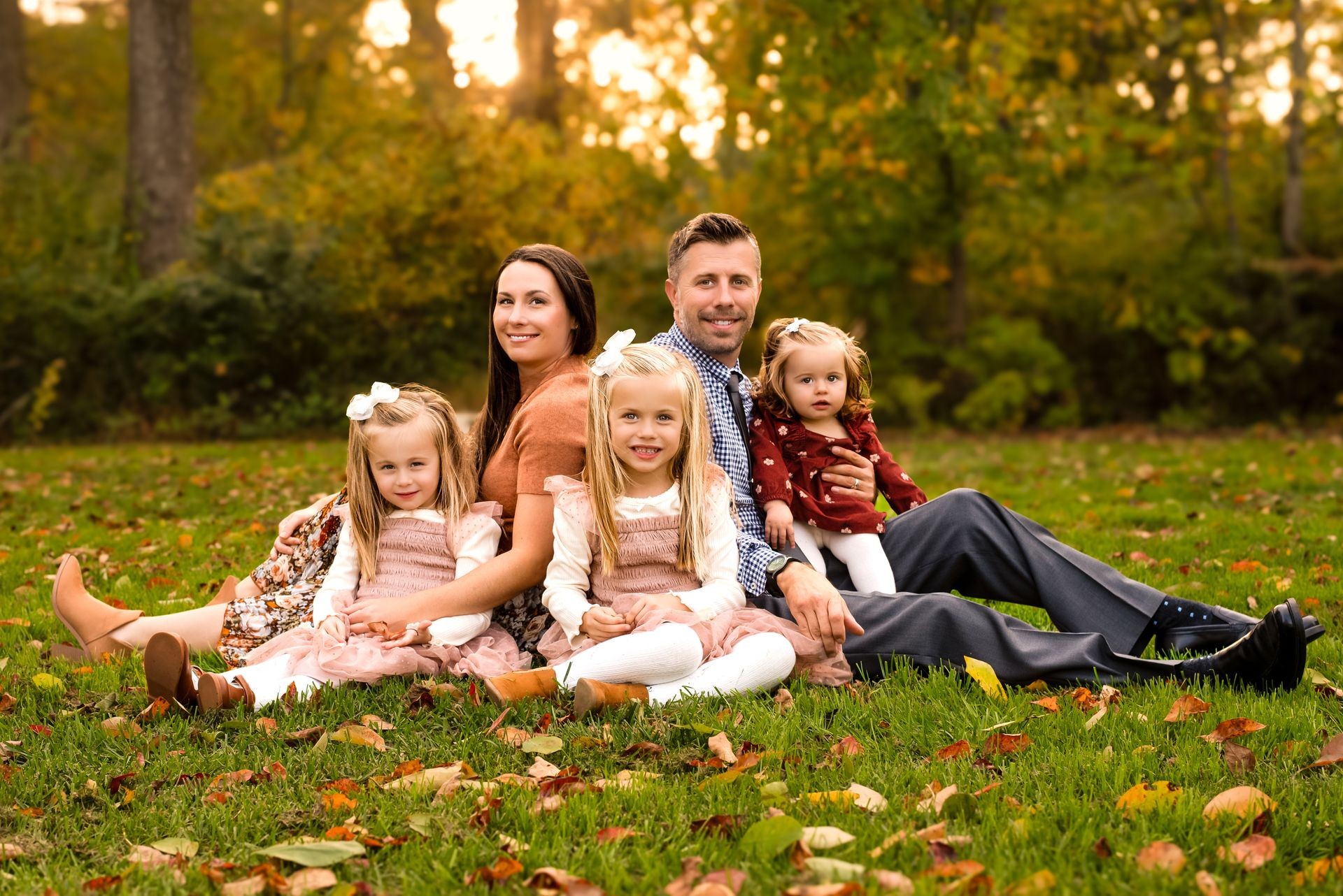 Family of five sitting on green grass surrounded by colorful fall trees at sunset. Parents smile beside their three young daughters, ages 1, 4, and 5, with the two older girls wearing matching fall outfits. Warm golden light shines through the trees, creating a cozy autumn atmosphere.