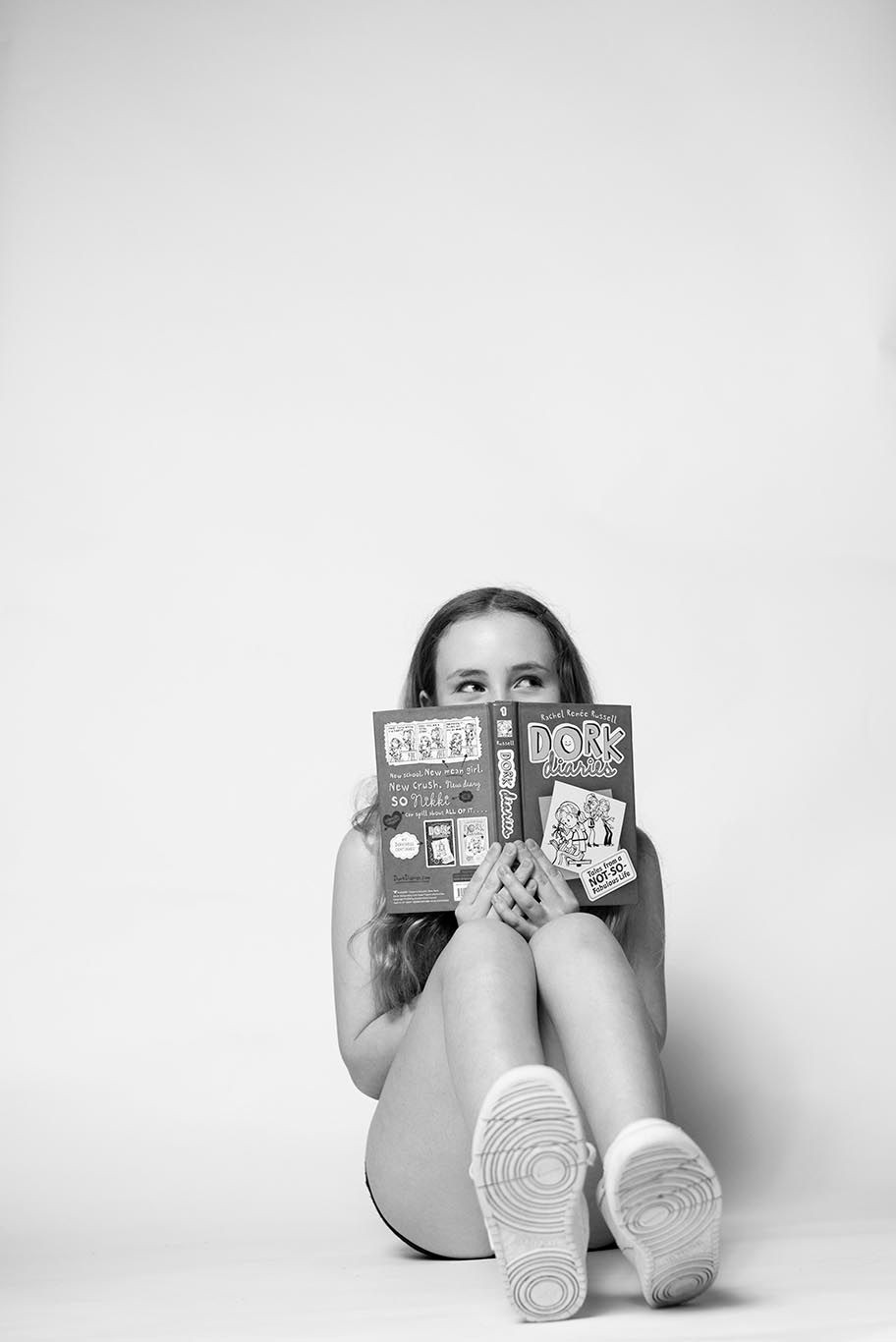 Person sitting on floor holding a Dork Diaries book in front of face, wearing sneakers.
