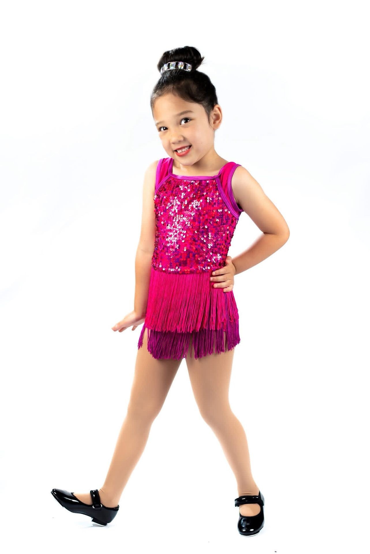 Young girl in sparkly pink dance costume and black shoes posing confidently against a white background.