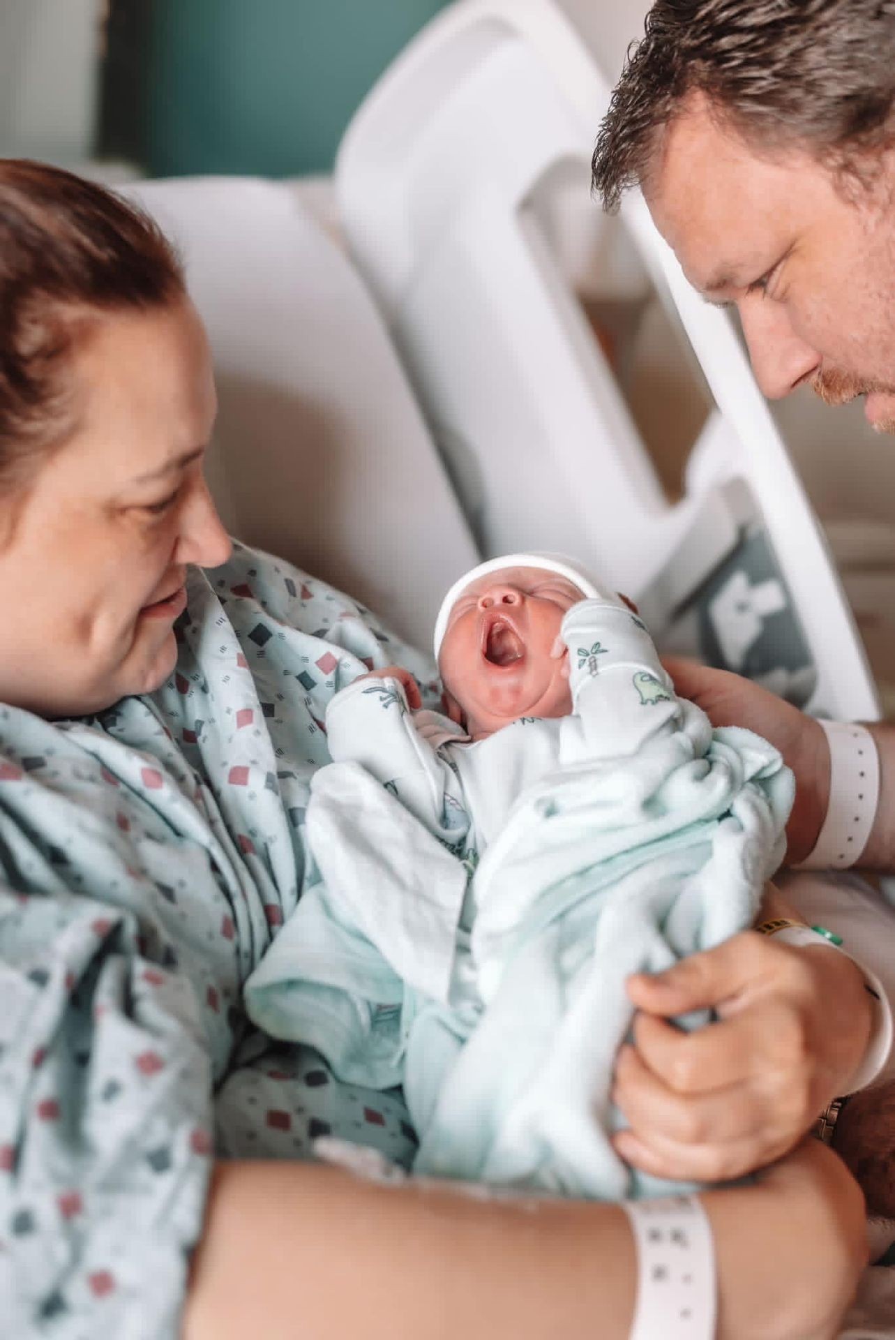 Newborn baby yawning while being held by parents in a hospital room.