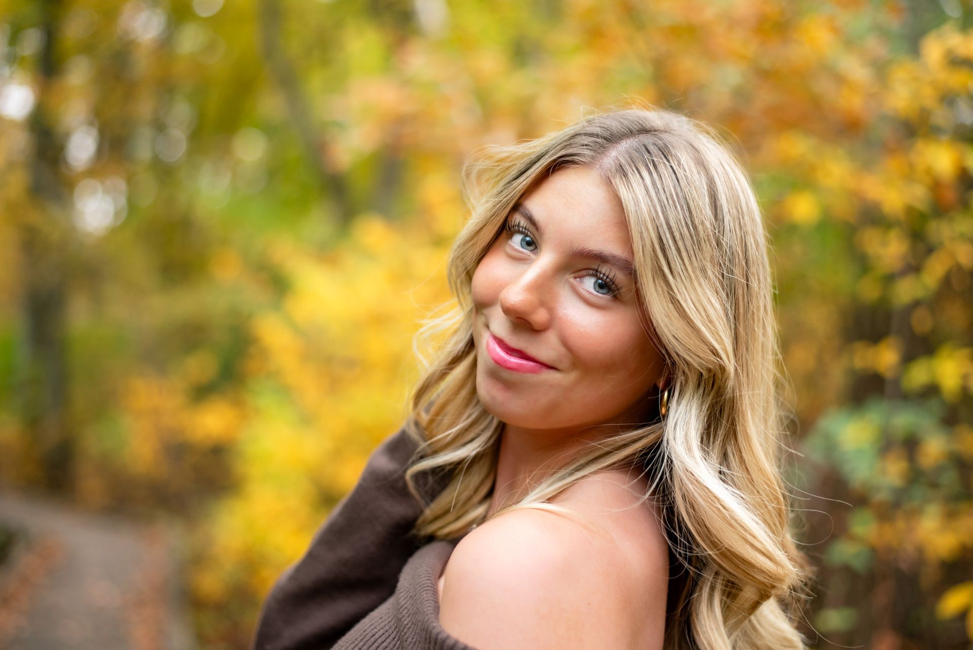 Woman with blonde hair smiling in a forest with yellow autumn leaves in the background.